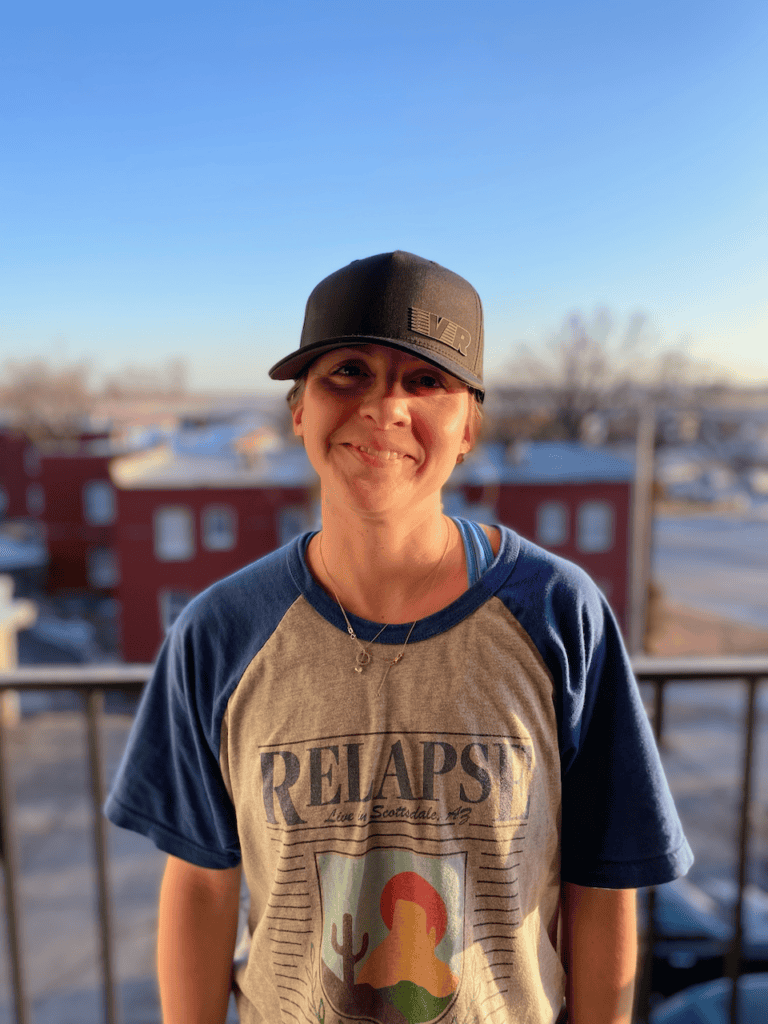 Smiling person on balcony, wearing a baseball cap.